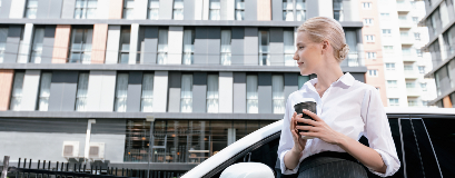 Eine Geschäftsfrau steht mit einem Kaffee in der Hand neben einem Auto im städtischen Umfeld.